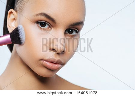 Cosmetic brushes. Pleasant charming nice woman holding a make up brush and applying face powder on her cheek while looking at you