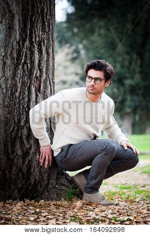 Handsome young man alone in nature near a tree outdoors. A charming young man with eyeglasses. The beautiful boy squatting on the ground leaning the tree. He dressing gowns autumn or winter. White sweater and gray trousers.