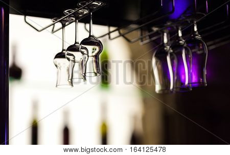 Glasses for grappa and tropical cocktails hang over a bar counter at restaurant against the background of the shelf with indistinct outlines of bottles.