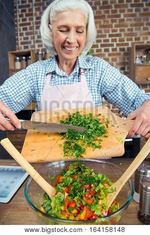 Smiling senior woman cutting salad greens at kitchen
