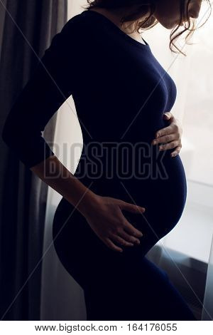 young pregnant girl in a blue bodycon dress standing at the window of the house