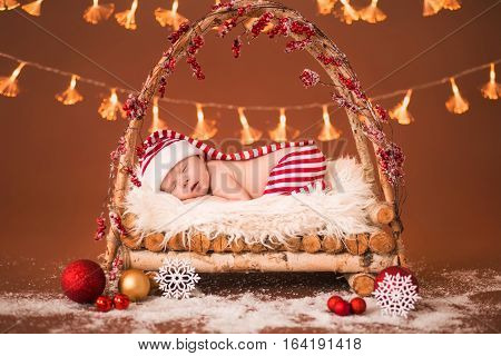 Newborn Baby Sleeping In A Wooden Crib. The Striped Suit Elf On A Brown Background.