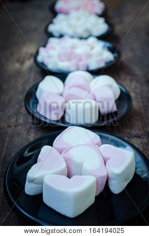 Colorful mini marshmallows in a black plates on a wooden background. Different mini white pink and orange puffy marshmallows. Marshmallow concept.