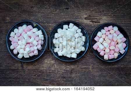 Colorful mini marshmallows in a black plates on a wooden background. Different mini white pink and orange puffy marshmallows. Marshmallow concept. Top view.