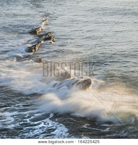 Wave Crashing Over Rocks In Ocean At Sunrise With Long Exposure