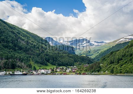 Small norwegian town and ferry harbor under the mountains with snowy peaks. View from the water landscape.