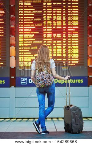 Tourist Girl With Backpack And Carry On Luggage In International Airport, Near Flight Information Bo