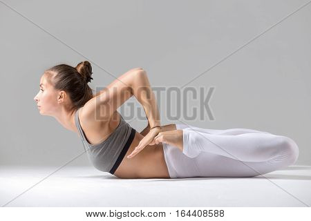 Young attractive woman practicing yoga, stretching in Frog exercise, Bhekasana pose, working out wearing sportswear, white pants, bra, indoor full length, isolated against grey studio background