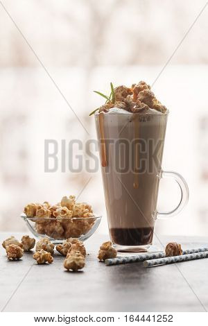 Glass of coffee latte with whipped cream, caramel popcorn, cinnamon and chocolate near the window