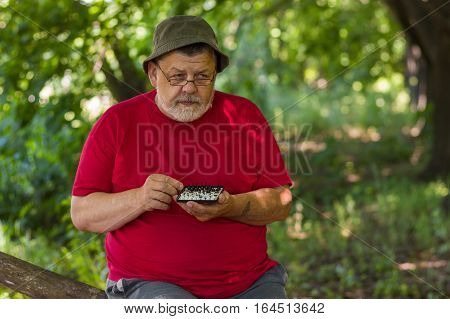 Bearded senior man wearing red t-shirt thinks over next move while playing mini-chess with himself under tree shadow in a summer park