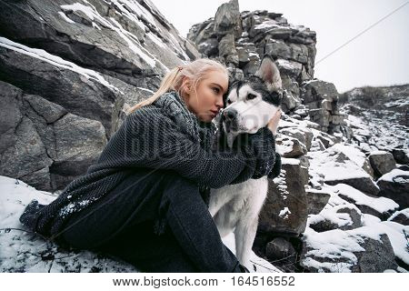 Girl with dog Malamute among rocks in winter. They sit on rocks among scattering of stones and snow. Girl hugs a dog's neck.