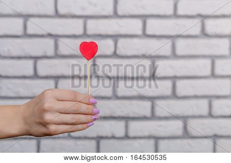 Woman's hand holding red heart shape on stick. White brick wall baskground. Valentine concept