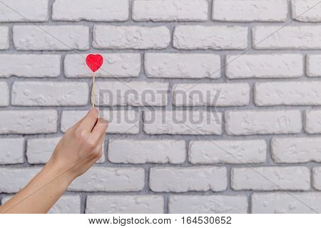 Woman's hand holding red heart shape on stick. White brick wall baskground. Valentine concept
