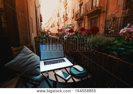 Cozy beautiful workspace on balcony with laptop smartphone wooden fence and a lot of flowers around on sunny summer day in historical district of Barcelona - El Born with facades of houses behind