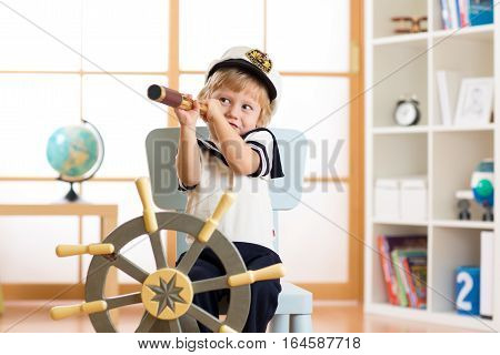 Kid dressed as a captain or sailor plays on chair as ship in his room. Child looks through telescope.