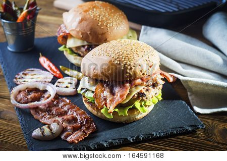 Fresh tasty burger. Close-up of two homemade burgers sesame buns with succulent beef patties grilled onions bacon and fresh salad served on black slate board on a rustic wood table.