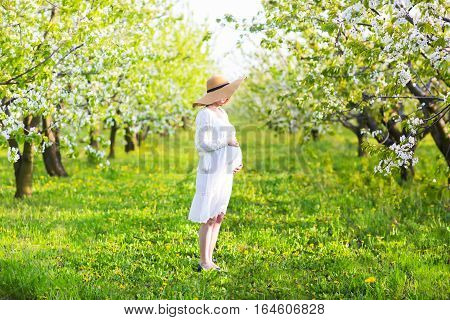 Beautiful pregnant woman wearing big hat and white dress in blooming garden