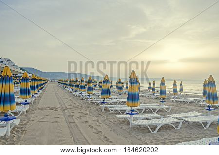 The Black Sea Shore At Sunrise, Beach Sand With Umbrellas And Sunbeds, Clouds And Blue Water.