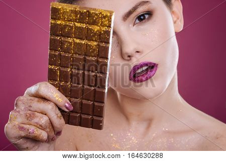 Sweet memories. Pleasant professioanl model holdign chocolate bar covered with tinsel while posing against pink background