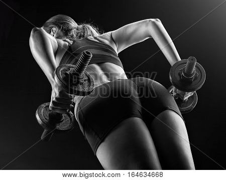 Focus on black shorts. Dark contrast image of fitness woman's back and buttocks. She is training - doing squats with dumbbells on black background in studio.