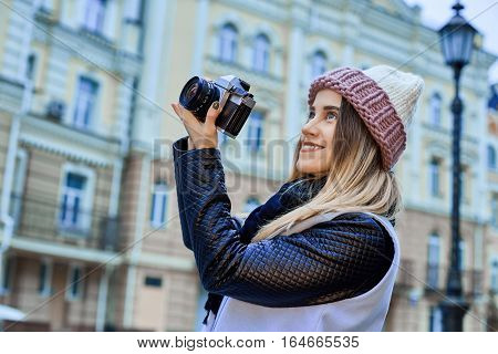 Beautiful girl photographer photographing the old town with retro camera. She is wearing a knitted cap