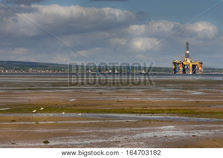 Cromarty Firth Scotland - June 3 2012: Panorama shot of Cromarty Firth at low tide with huge retired and damaged oil drill platform. Wide skies. Green hills. Brown marsh land.
