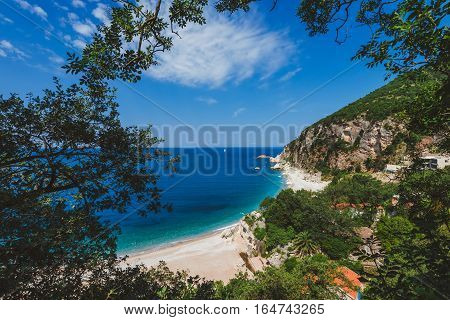 Adriatic sea coast by sunny day summer landscape. Pebble beach, house roofs and green tree brunches under blue sky near Perazica Do village on the way to Petrovac, Montenegro.