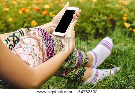 close up white mobile phone in fashionable trendy woman hands on the background of green grass with flowers during an exercise break