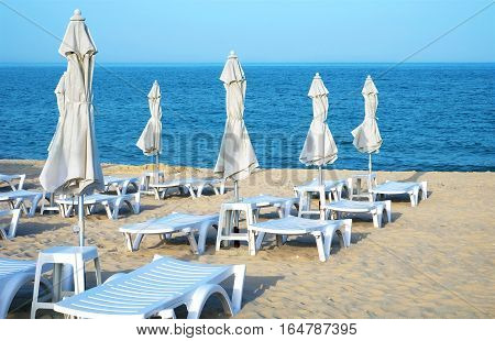 Empty sun loungers and closed umbrellas on a deserted sea sandy beach on a clear day.