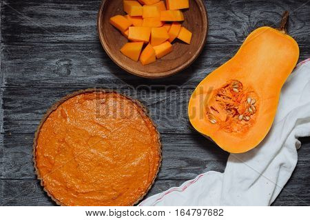 Overhead shot of pumpkin pie half of butternut squash with seeds and pumpkin pieces in clay bowl on background of rustic gray wood planks. Homemade pie cooking in autumn healthy food.