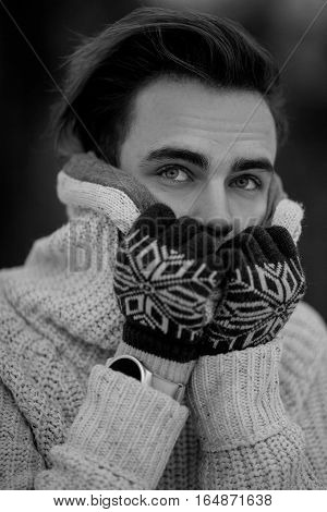 Young man portrait in cold winter weather. He is cold and he wrapped himself in sweater put on gloves. Black and white image.