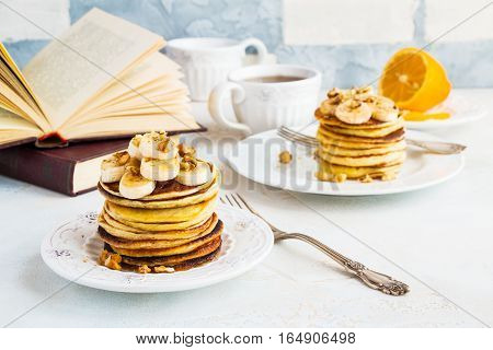 Stack of homemade pancakes with banana maple syrup and walnuts on vintage plate. Fork fresh sliced fresh lemon cup of tea open book white and gray concrete background.