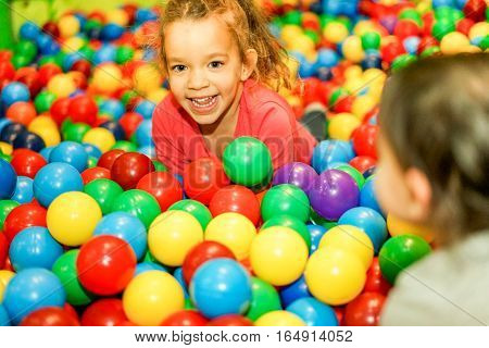 Cheerful children playing inside ball pit swimming pool - Happy people having fun in baby playground indoor - Childhood and friendship concept - Soft focus on left female face - Warm vivid filter