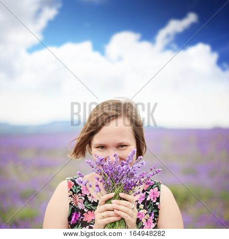 Young beautiful smiling woman in blouse with flower ornament standing at the background of purple flower field with bouquet of lavender in hands.