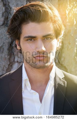 Model handsome Italian elegant man. Intense outdoor light. Portrait of a handsome young man. The boy is outdoors near a tree. He is watching with calm and peaceful attitude. He wears stylish elegant clothes. Warm light background.