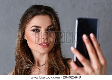 Beautiful European Woman Portrait. Worth A Gray Background Uses A Smartphone Makes Selfie And Reads 