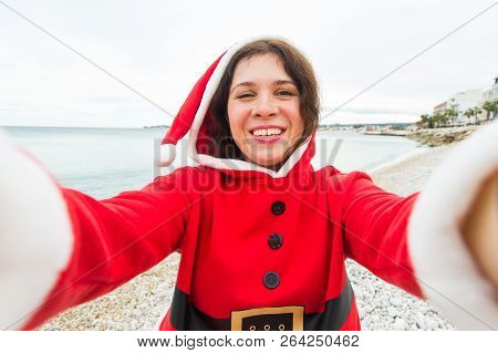 Christmas, Humor And People Concept - Young Woman In Santa Suit Making Selfie Near The Sea At Summer