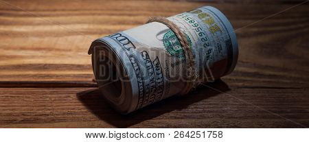 A Roll Of Dollars On A Wooden Textured Table In The Dark Illuminated By A Ray Of Light.