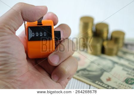 Hand Holding Tally Counter Or Counting Machine With 0000 Number, Coin Stack And Money On White Woode