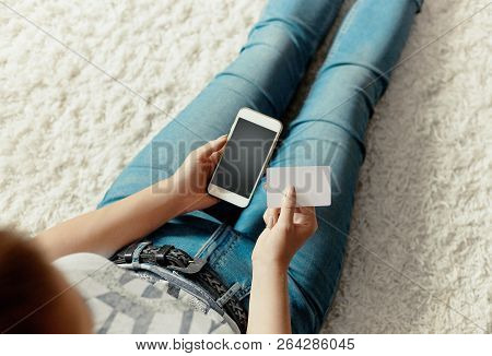 Young Woman Holding Credit Card And Using Phone Sitting On Floor. Payment.  Online Shopping Concept.