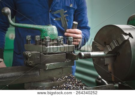 Turner Is Working On A Lathe Machine In A Factory. Worker Is Measuring A Produced Detail By Caliper.