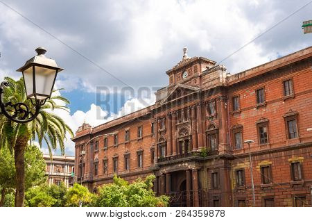 Оld Red Brown Brick Building With Clock Watch, Street Lamp Lights And Palm Trees In Front Of Cloudy 