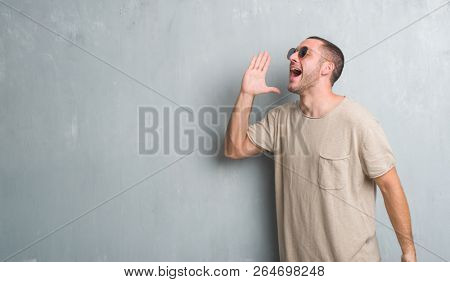 Young caucasian man over grey grunge wall wearing sunglasses shouting and screaming loud to side with hand on mouth. Communication concept.