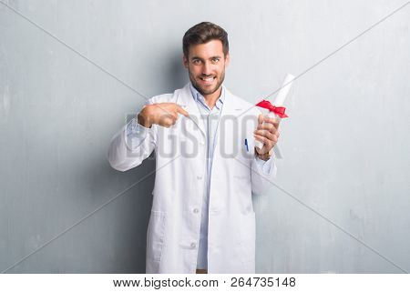 Handsome young doctor man over grey grunge wall holding diploma with surprise face pointing finger to himself