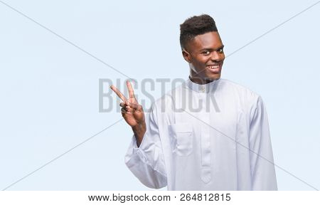 Young arabic african man wearing traditional djellaba over isolated background smiling with happy face winking at the camera doing victory sign. Number two.
