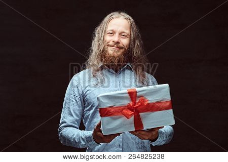 Happy Male With Long Hair And Beard In Blue Shirt Holds Gift Box. Isolated On A Dark Background.