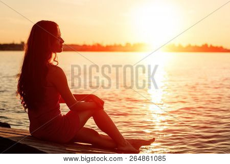 Romantic girl sitting on the pier near the water at sunset. Woman by the sea. Model on the beach on a background of sunlight. Siluet in backlight.