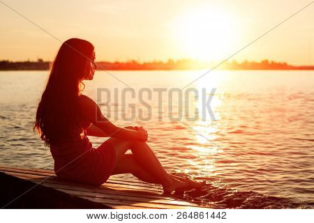 Romantic girl sitting on the pier near the water at sunset. Woman by the sea. Model on the beach on a background of sunlight. Siluet in backlight.