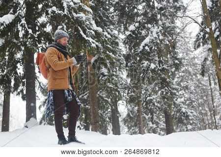 Serious young male hiker in warm coat standing in winter forest and using gadget while finding trail via gps navigation