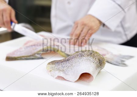 A Large Zander Fish Fillet Lying On A White Chopping Board. A Chef Filleting A Fish In The Backgroun